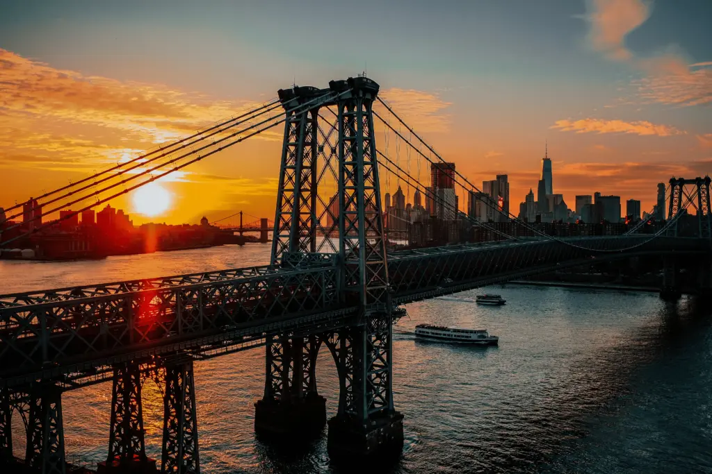 Vista da estrutura de uma grande ponte suspensa em Nova York cruzando o rio durante um pôr do sol alaranjado, com silhuetas de prédios ao fundo e barcos na água.