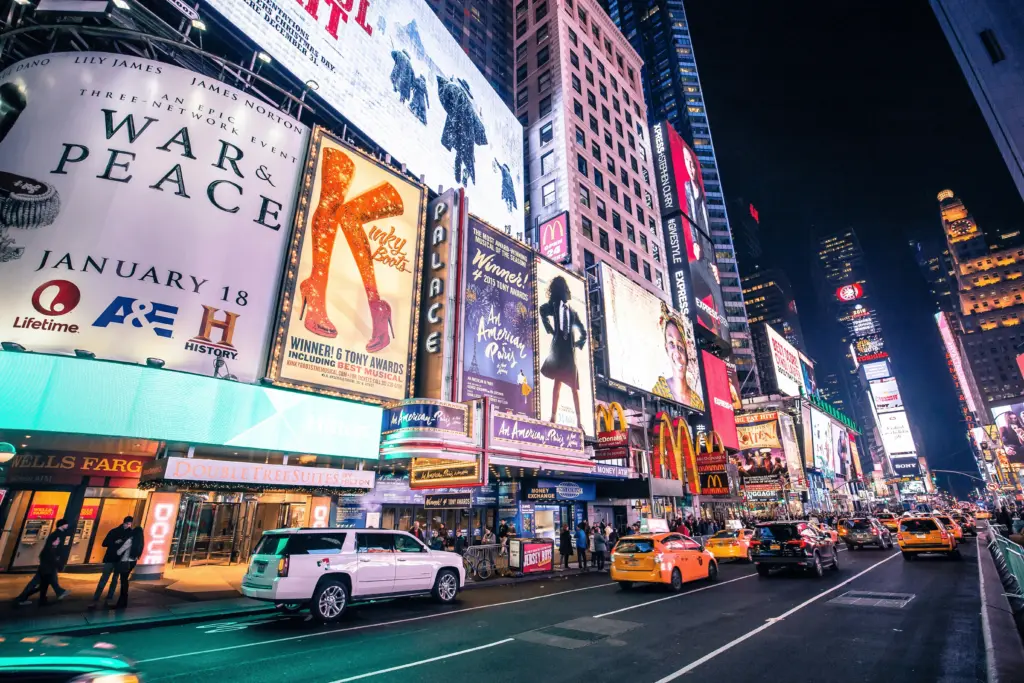 Vista noturna da Times Square em Nova York iluminada por grandes painéis publicitários luminosos, letreiros de teatros da Broadway e movimentação de táxis amarelos na avenida.