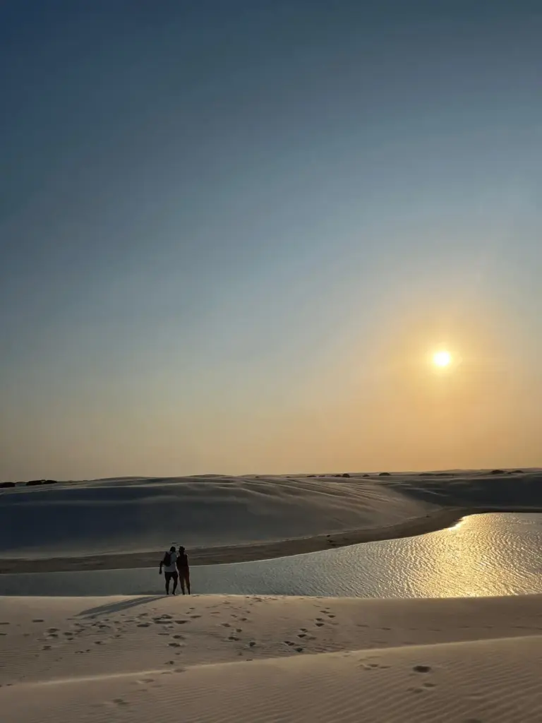 Foto original da KG Viagens retratando um casal admirando o pôr do sol nos Lençóis Maranhenses. As lagoas cristalinas e as dunas brancas criam uma experiência inesquecível para quem visita este paraíso natural.